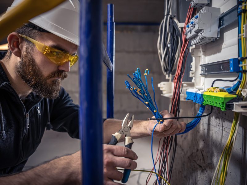 A male electrician works in a switchboard with an electrical connecting cable, connects the equipment with tools.